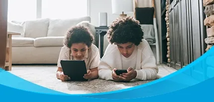 Two curly-haired kids lie on a living room rug using a tablet and phone.